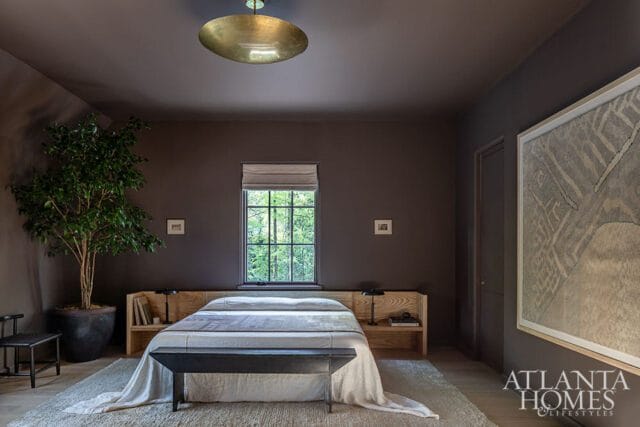 Dark gray bedroom with a queen-size bed featuring a light wood headboard with built-in shelves, a white duvet, and a dark bench at the foot. A large potted tree sits to the left, and a framed abstract artwork hangs on the right wall.