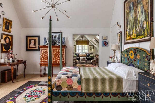 Bedroom with a four-poster bed featuring a colorful, patterned headboard and a patchwork quilt. Artwork adorns the walls, and a doorway reveals a glimpse of a living area.