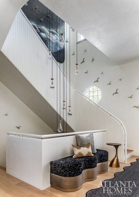 Foyer with a curved staircase, white paneled walls, and a large abstract mural. A series of glass pendant lights hang down the stairwell. Decorative bird sculptures are mounted on the walls.