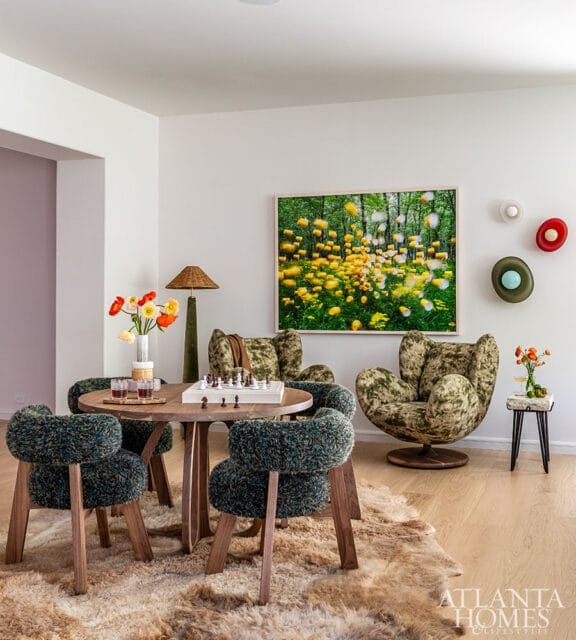 Game room with round wood table, four textured green chairs, two plush armchairs, and a large abstract painting of yellow flowers in a forest.