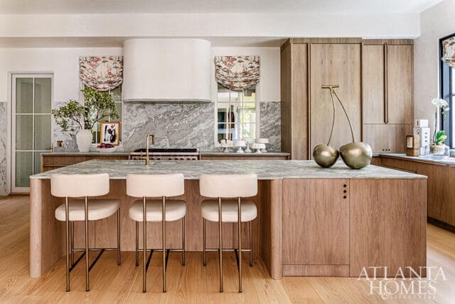 Modern kitchen with a large marble island, light wood cabinetry, and cream upholstered bar stools. A white range hood is centered above a marble backsplash.