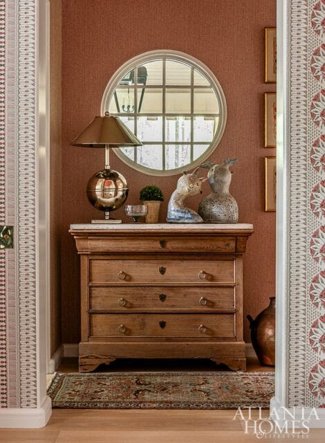 Mudroom with a wooden dresser topped with a marble slab, a globe lamp, decorative animal sculptures, and a round window with a grid pattern. The walls are covered in textured rust-colored wallpaper and patterned wallpaper borders.