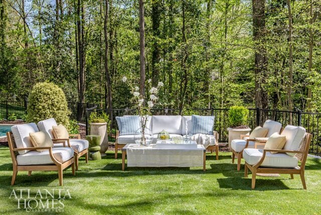 Teak outdoor furniture with white cushions and blue striped pillows arranged on a green lawn, backed by a dense forest and black metal fence.