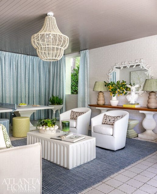 Living room with white swivel chairs, fluted white coffee table, blue rug, and light blue patterned curtains. A large woven chandelier hangs from the gray wood-paneled ceiling.