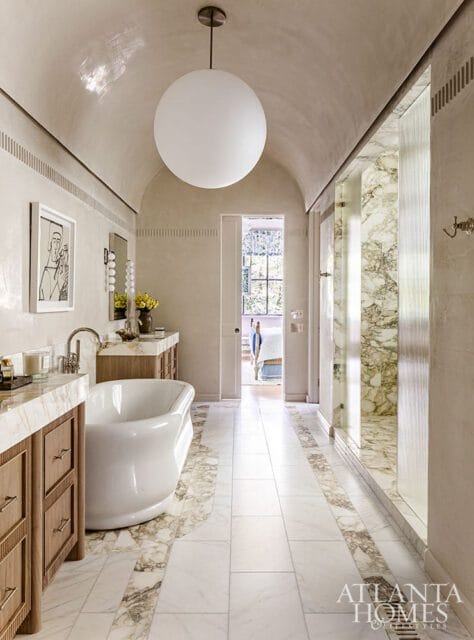 Primary bathroom with a freestanding tub, marble countertops, wood vanities, and a marble-tiled shower. A large spherical pendant light hangs from the vaulted ceiling.