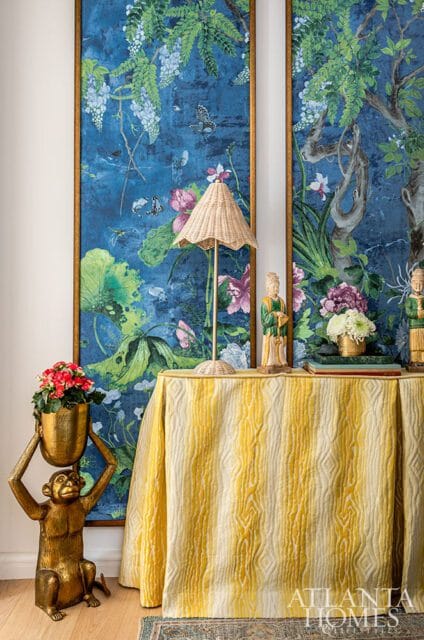 Primary bedroom with a gold monkey statue holding a planter, a table draped in a yellow and cream striped cloth, and two framed botanical murals featuring blue backgrounds, lush greenery, and butterflies.