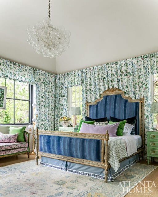 Primary bedroom with a blue upholstered bed, floral curtains, green nightstands, and a patterned rug. A glass chandelier hangs from the ceiling.