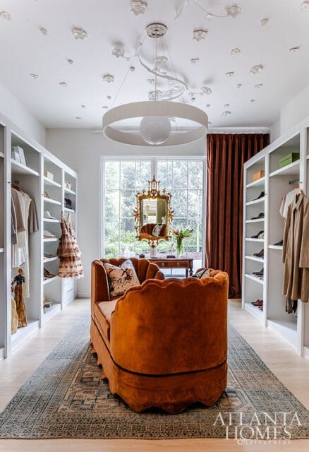 Walk-in closet with white built-in shelving, an orange velvet scalloped sofa, a patterned rug, and a large window with brown curtains.