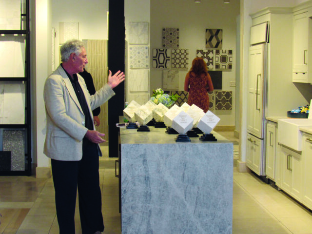 Man in blazer gesturing near marble island with awards and tile samples on wall