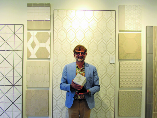 Man in blue jacket holding award in front of patterned wallpaper and tile samples