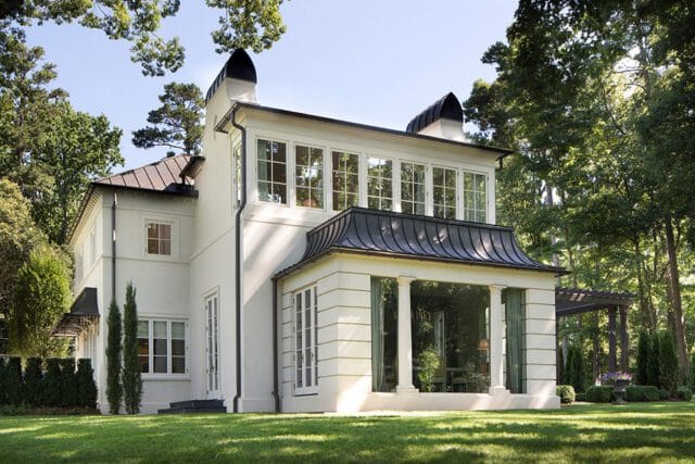 White house with black metal roof, columns, and large windows surrounded by trees.