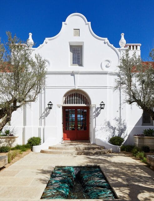 White stucco house with red double doors and a decorative water feature in the foreground.