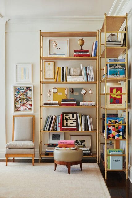 Gold bookshelves filled with books, art, and decor. A chair and ottoman are nearby.