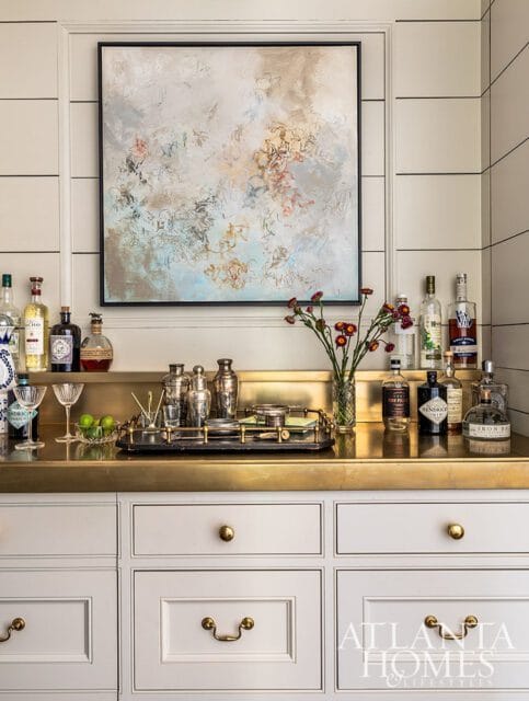 Bar area with brass countertop, white cabinetry, abstract art, and liquor bottles.