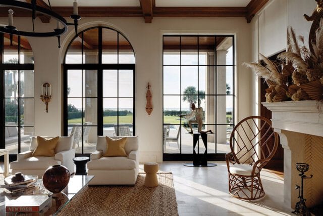 Living room with white chairs, arched windows, and fireplace. View of ocean and palm trees.