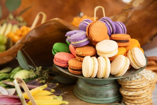 Pile of colorful macarons on a green pedestal stand with crackers and vegetables.