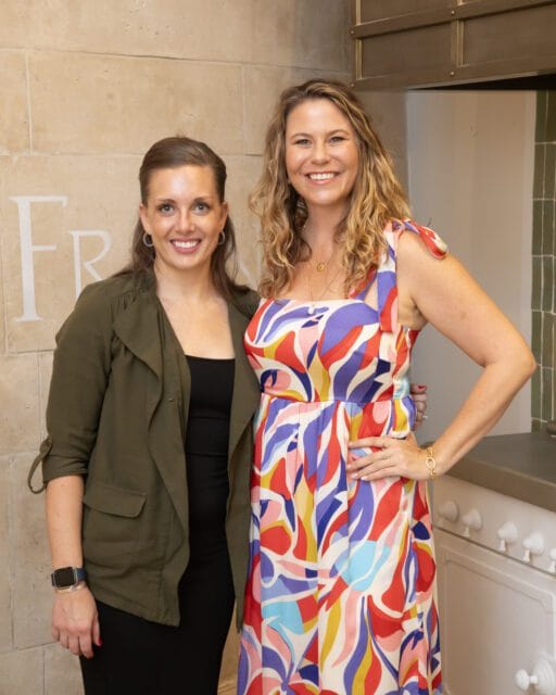 Two women smiling, one in a green jacket and black dress, the other in a colorful abstract print