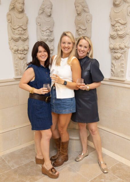Three women smiling, two holding drinks, with stone sculptures on the wall.