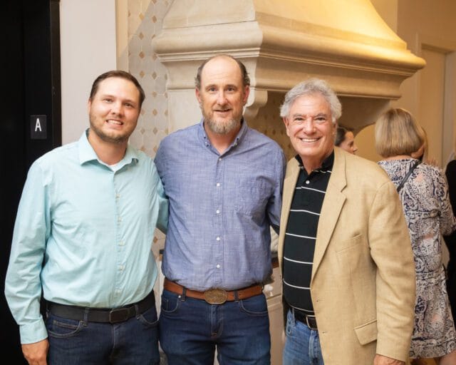 Three men smiling, with a decorative stone fireplace behind them.