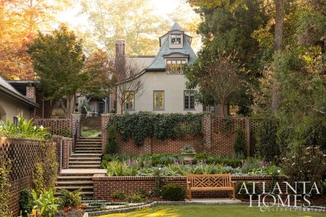 French-style house with gray slate roof, brick accents, and tiered garden with bench.