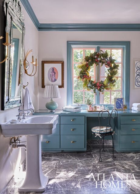Blue vanity with gold hardware, pedestal sink, and marble floor. Wreath on window.