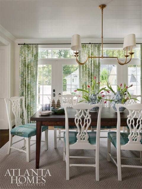 Dining room with dark wood table, white chairs with plaid seats, green floral curtains, and brass
