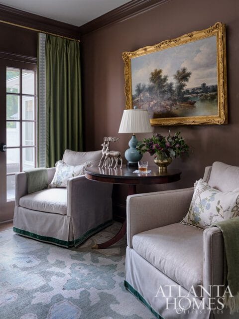 Two armchairs with green trim flank a round table with a lamp, deer figurines, and a painting.