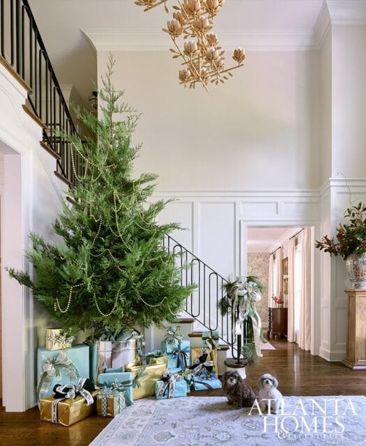 Foyer decorated for Christmas with a tall evergreen tree adorned with pearl garland, surrounded by wrapped gifts. Two dogs sit on a patterned rug near a staircase with a black railing.