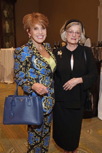 Woman in patterned suit with blue handbag and woman in black suit with brooch