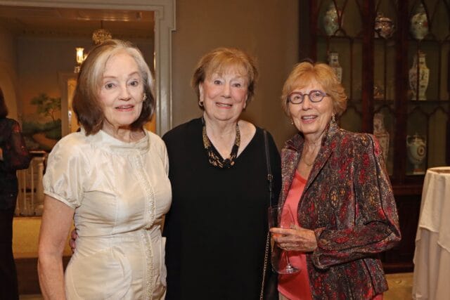Three women posing for a photo. The woman on the left wears a cream dress. The woman in the middle