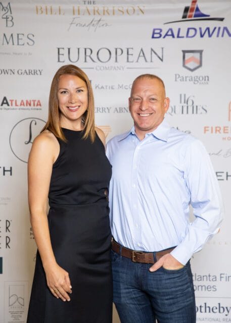 Man in blue dress shirt and woman in black dress smiling, standing in front of a sponsor banner
