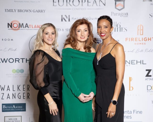 Three women posing in front of a sponsor wall at an event.