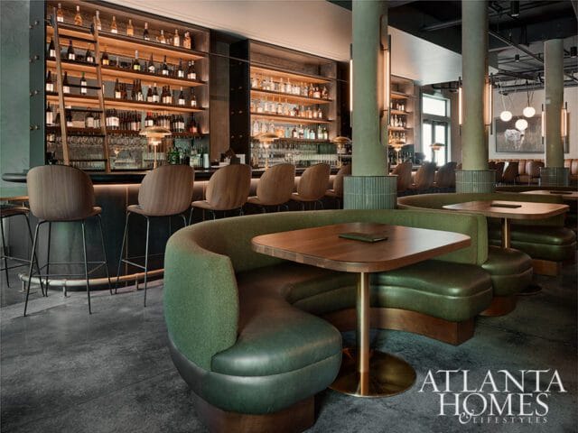 Green banquette seating and wood tables in a restaurant with a bar and shelves of liquor bottles.