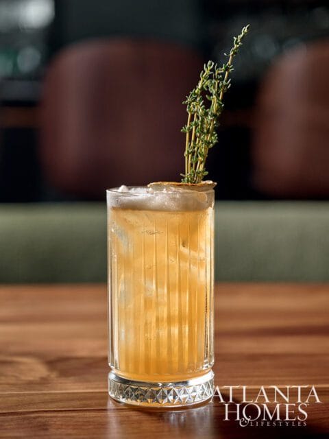 Tall glass of amber cocktail with ice, garnished with herbs and a citrus slice.