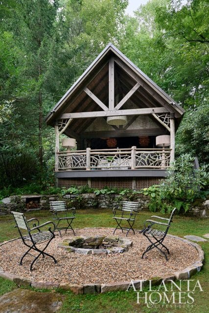Rustic wooden pavilion with stone fire pit and seating in a garden setting.