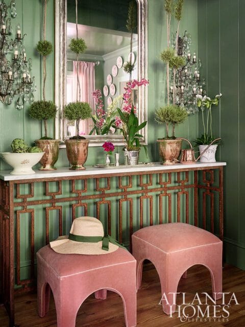 Green paneled dining room with marble console table, pink ottomans, and topiaries.