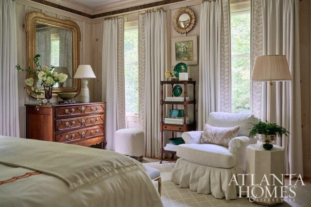 Primary bedroom with antique dresser, white armchair, and Atlanta Homes & Lifestyles logo.