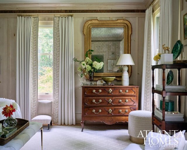 Primary bedroom with wood paneled walls, ornate mirror, antique dresser, and floral curtains.