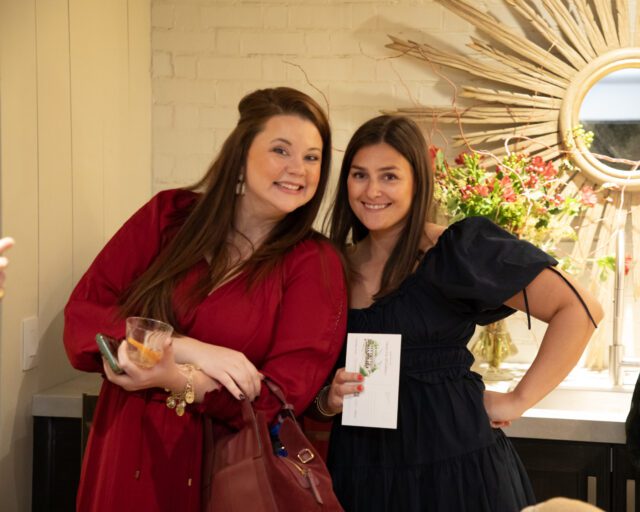 Two women smiling, one in a red dress holding a drink, the other in a navy dress holding a card.