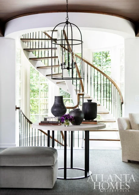 Round table with black vases and purple flowers, near a curved staircase and ottoman.