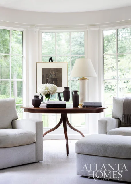 Light gray armchairs and ottoman in a room with a bay window, round wooden table with vases and