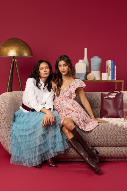 Two women sitting on a textured sofa against a red backdrop. One wears a white shirt and blue