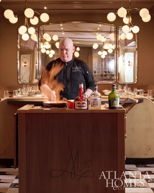 Chef cooking with flames in a restaurant with a mirrored wall and globe pendant lights.