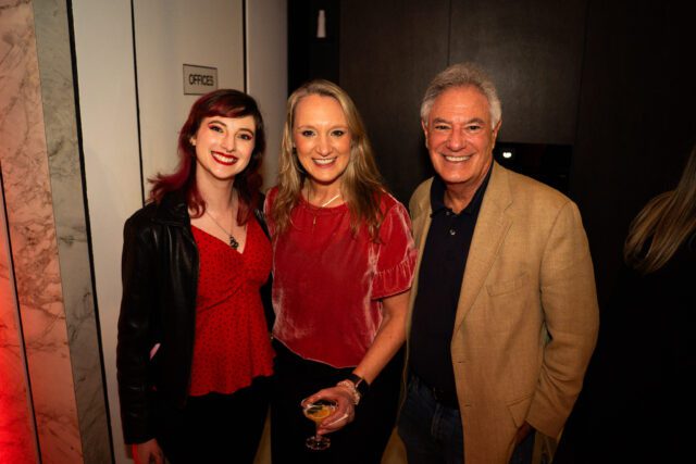 Three people smiling, two women and one man. The woman on the left has red hair and a black leather