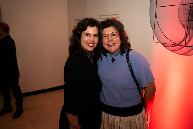 Two women smiling at the camera. The woman on the left wears a black top and the woman on the right