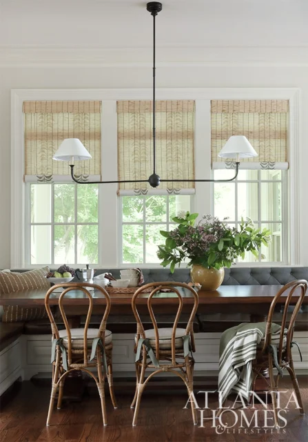 Dining nook with tufted banquette, wooden table, rattan chairs, and bamboo blinds.