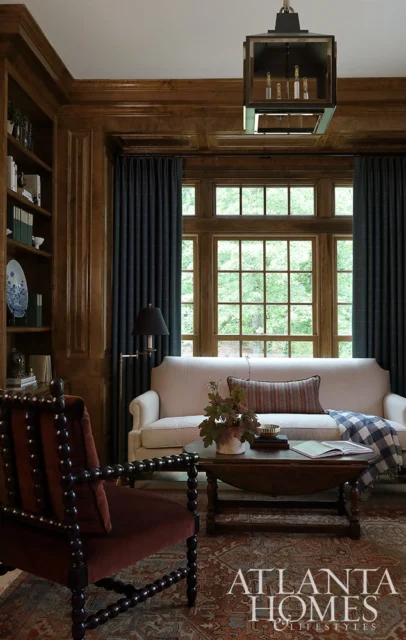 Living room with wood paneling, cream sofa, dark curtains, and a large window overlooking trees.