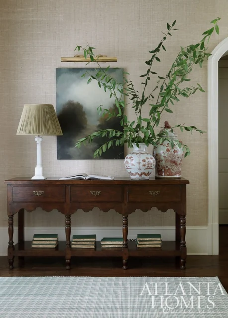Dark wood console table with a lamp, painting, and ginger jars. Grasscloth wallpaper.