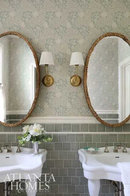 Bathroom with floral wallpaper, two oval mirrors, brass sconces, and double sinks with green tile.