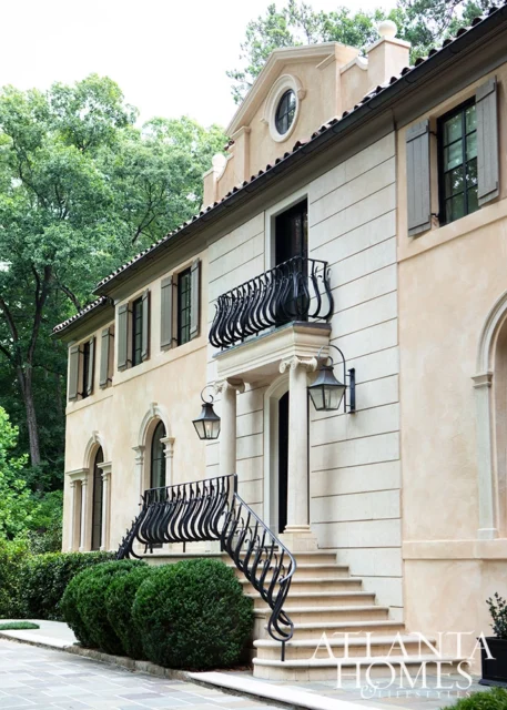 Light-colored stucco house with black wrought iron balcony and stairs, surrounded by trees.